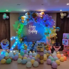 A vibrant Baby Shark themed birthday party. The decoration features a multi-colored balloon arch, a "Happy Birthday" neon sign, and character cutouts, creating an underwater feel.