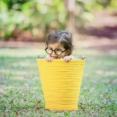 A playful outdoor shot of a curious baby with glasses peeking out of a bright yellow bucket. This type of fun and creative kids' portrait captures their charming personality.