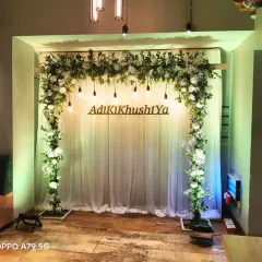 A fresh and simple engagement backdrop with a white floral and greenery arch. The couple's hashtag, "AdiKiKhushiYa," is displayed in the center for a personal touch.