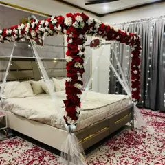 A classic wedding night room decoration. The bed is framed by a four-poster structure covered in red and white flowers, with delicate white drapes and fairy lights adding to the romance.