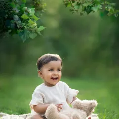 A baby laughs with delight while playing with a teddy bear during an outdoor photoshoot. The golden light filtering through the trees is just magical.