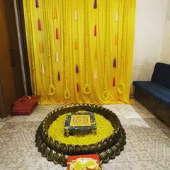 For a simple home Haldi function, we can create a beautiful setup with a yellow curtain backdrop and a traditional brass urli filled with marigold petals for the ceremony.