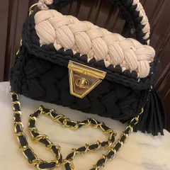 A close-up of the black and beige crochet bag, highlighting the braided texture and the secure gold-tone clasp. The tassel is also handmade with the same yarn.