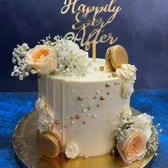 A classic white drip cake for a 'Happily Ever After' moment. This elegant design is adorned with fresh peach roses, baby's breath, and shimmering gold macarons, perfect for engagements, anniversaries, or intimate weddings.