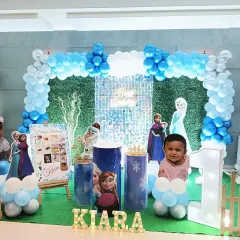 A Frozen themed first birthday party featuring a greenery and shimmer backdrop, a balloon arch in shades of blue, and cutouts of Anna, Elsa, and Olaf.