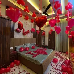A full room decorated for a romantic surprise. The setup includes red heart-shaped balloons on the ceiling, "LOVE" foil letters, and "I ❤️ U" spelled out with rose petals on the bed.