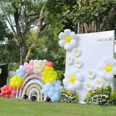 A stunning photo backdrop for a sunshine-themed party. This setup features a large white panel adorned with giant balloon daisies and a pastel rainbow arch made of wood and balloons.