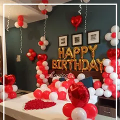 A full view of a romantic red and white room decoration. The bed is covered in balloons and rose petals, with heart-shaped foil balloons and 'Happy Birthday' letters on the wall.