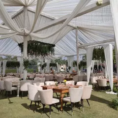 A chic lounge seating arrangement for a daytime outdoor event. The combination of modern upholstered chairs, wooden tables, and draped white canopies creates a comfortable and stylish space for guests to relax.