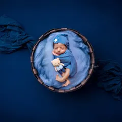 A classic newborn pose, with the baby swaddled snugly in blue and tucked into a rustic basket with a tiny teddy bear. The deep blue background creates a rich, portrait-like effect.