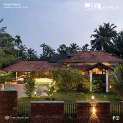 An evening view of Kuttiyil House, highlighting the interplay of light and structure. The design uses multiple sloped roofs characteristic of Kerala architecture and low-level landscape lighting to accentuate the natural textures of the laterite stone walls and surrounding greenery.