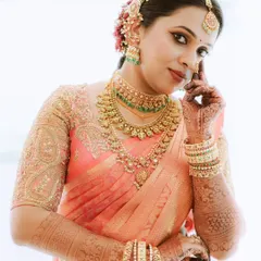 A detailed portrait of a bride in a peach-colored saree, highlighting her intricate temple jewellery and henna-adorned hands.