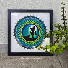 Another shot of the Krishna and calf mandala, placed next to a plant. The natural light highlights the vibrant blues and greens in the artwork.