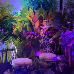 A lush and moody photo booth from our jungle-themed event. A vibrant tropical wallpaper serves as the backdrop, with real foliage and dramatic purple and blue lighting creating a mysterious, wild paradise for guests to pose in.