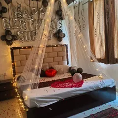 A surprise birthday room decoration with a canopy tent over the bed. The black and silver balloons, fairy lights, and rose petals create a stylish and romantic atmosphere.