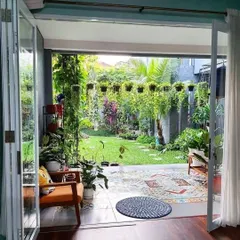A view from inside looking out to a cozy patio and garden. The design uses patterned tiles on the patio floor, comfortable seating, and a variety of hanging and potted plants to create a seamless indoor-outdoor living experience.