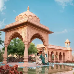 A wide-angle shot showcasing the grandeur of the rajwada location. The couple is seated by the water, framed by the majestic pavilion, creating a breathtaking image that looks like a scene from a movie.