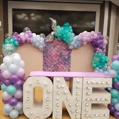 Celebrating a first birthday is a huge milestone. This setup features large light-up "ONE" marquee letters, a mermaid-scale backdrop, and a balloon arch in mermaid-inspired colors of purple, teal, and lavender.