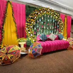 A full view of a traditional Mehendi seating area. The combination of a bright pink sofa, a floral arch of marigolds, and colorful Rajasthani umbrellas creates a picture-perfect spot for the bride.
