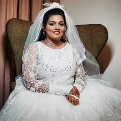 The bride seated, looking calm and beautiful before her ceremony. The full white gown and veil are the stars, supported by a polished and timeless makeup and hair look.