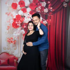 An expecting couple shares a loving embrace in front of a bold red floral backdrop, with the mother-to-be in a classic black dress.