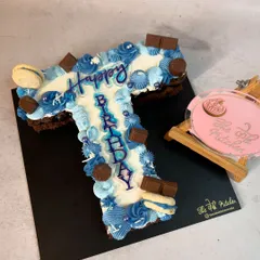 A monogram cake in the shape of the letter "T," decorated in shades of blue with buttercream piping, chocolate pieces, and macarons.
