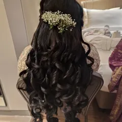 A romantic half-up hairstyle with voluminous, structured curls perfect for an engagement or sangeet. The look is finished with a sprig of fresh baby's breath, adding a touch of softness and elegance.