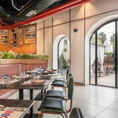 The main dining area at Burosu features long pink leather-upholstered booths and terrazzo tabletops. The arched doorways lead to an outdoor seating area, creating a seamless flow between the indoor and outdoor spaces.