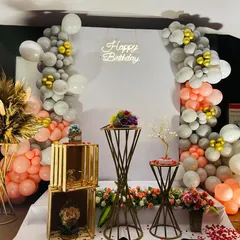 An elegant birthday decoration featuring a sophisticated color palette of grey, peach, and gold. The setup includes two asymmetrical balloon garlands framing a central backdrop with a 'Happy Birthday' neon sign.