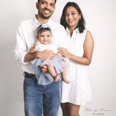 A classic studio family portrait. The parents and their baby girl are dressed in coordinating white and blue, smiling together against a clean white background for a timeless look.