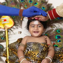 A joyful and divine baby photoshoot. This little one is styled as Lord Murugan, with peacock feathers and a Trishula, blessed by his parents' hands in a creative and heartwarming setup.