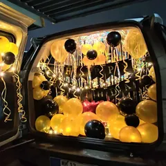 This car trunk decoration features yellow and black balloons illuminated by warm fairy lights. A 'Happy Birthday' banner is strung across the back, creating a glowing and cheerful surprise.