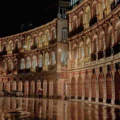 A lone figure with a red umbrella walks along a rain-slicked street at night, with the illuminated arches of a colonial-era building reflected on the wet pavement. This is a classic, atmospheric Mumbai monsoon moment.