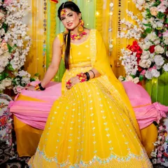 A happy bride poses at her Haldi ceremony. The backdrop is a cheerful mix of yellow drapes, marigold strings, and a large floral arch, perfectly complementing her bright yellow lehenga.