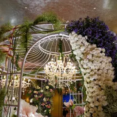A beautiful white birdcage-style gazebo, adorned with cascading flowers in shades of purple and white. The crystal chandelier inside adds a touch of classic elegance to the whimsical structure.