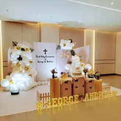 For George and Andrea's First Holy Communion, we designed a sophisticated and modern stage. The decor combines clean white panels, elegant balloon clouds, and touches of gold, with personalized marquee lights to make the day truly special and blessed.