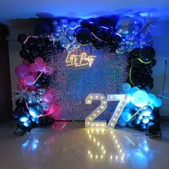 This setup for a 27th birthday party has a vibrant, fun feel. It features a silver sequin wall, a 'Let's Party' neon sign, marquee numbers, and colorful balloon clusters with blue and pink lighting.