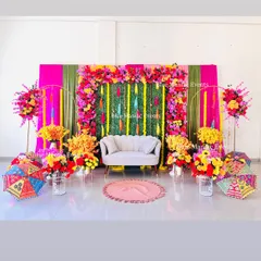 For a vibrant Mehendi or Haldi, I can transform any space with a burst of color. This grand setup uses a lush green wall as a base, framed by a riot of pink, yellow, and red artificial flowers, with traditional umbrellas and floral arrangements completing the festive look.