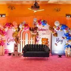 A fun 'Boy or Girl' themed baby shower stage setup. The backdrop is split into pink and blue sections, each with its own balloon arch, a teddy bear prop, and a white picket fence accent.