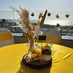 A close-up of a tabletop centerpiece for our bee-themed baby shower. This arrangement sits on a rustic wood slice and includes pampas grass, a cute bee cutout, yellow flowers, and a small chalkboard sign, all set against a beautiful seaside view.