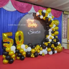 We designed this complete stage setup for a 50th birthday party in a hall. It features a custom circular backdrop with the name 'Sweta', a neon sign, large foil '50' balloons, and an extensive balloon garland in black, gold, and white.