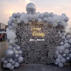 We take the party anywhere, even your rooftop. This setup features a brilliant silver shimmer wall framed by a white and silver balloon arch, creating a chic and elegant photo corner for an outdoor celebration.