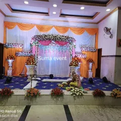 A traditional stage decoration for a naming ceremony in a function hall. The setup includes a central floral arrangement, decorative urns with flowers, and bright orange and white drapes.