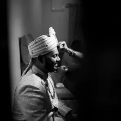 A candid black and white shot of the groom getting his turban adjusted, a quiet moment of preparation before the main ceremony.