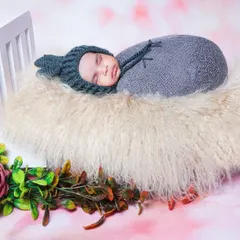 A newborn wearing a cute bunny-eared hat sleeps on a fluffy bed prop. The pink floral background adds a soft, dreamy quality to the image.