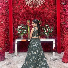 A happy client rocking a dark green floral print lehenga set against a stunning red flower wall. This shows how our outfits are perfect for any grand celebration.