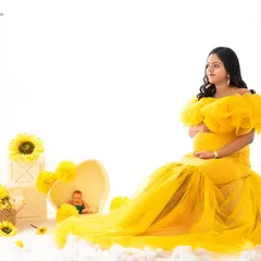 A bright and cheerful studio setup with a mom-to-be in a sunny yellow gown. The scene is decorated with sunflowers and white clouds, creating a whimsical and airy high-key maternity portrait.