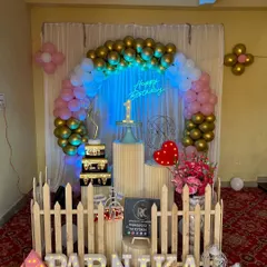 The white picket fence is a popular prop that adds a sweet, storybook touch to any celebration. Here it encloses a display area for the cake and gifts, personalized with the name 'PARNIKA' in light-up letters.
