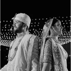 A strong, classic black and white portrait. The couple stands back-to-back against a backdrop of lights, symbolizing their partnership and individual strength.