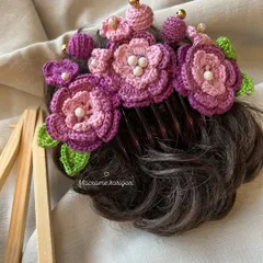 A close-up of the purple floral crochet bun comb, showing the intricate details of the petals and the pearl accents. It's designed to sit beautifully on a hair bun.